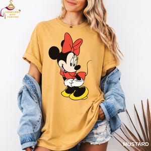 May include: Mustard-colored t-shirt featuring a graphic of Minnie Mouse in a red bow, dress, and shoes. The shirt has rolled-up sleeves and is worn with a denim jacket. The word "MUSTARD" is printed in the bottom right corner.