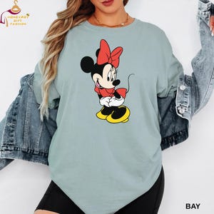 May include: A light blue t-shirt featuring a Minnie Mouse graphic. Minnie Mouse is depicted in her classic outfit: a red bow, red dress, white gloves, and yellow shoes. The shirt has the word "BAY" printed on the bottom right.