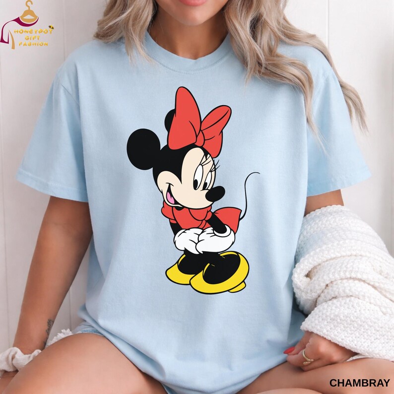 May include: Light blue t-shirt featuring a Minnie Mouse graphic. The cartoon character is depicted in a classic pose, wearing a red bow, scarf, and dress, with yellow shoes. The word "CHAMBRAY" is printed on the bottom right.