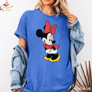 May include: A blue t-shirt featuring a graphic of Minnie Mouse in a red dress and bow, with yellow shoes. The shirt has rolled-up sleeves and the text "FLO BLUE" is visible in the bottom right corner.