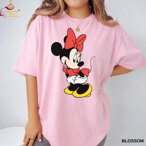 May include: Pink t-shirt featuring a Minnie Mouse graphic. The cartoon character is depicted in a red bow, dress, and shoes, with black ears and a smiling face. The word "BLOSSOM" is printed in white on the bottom right.