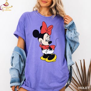 May include: Violet-colored t-shirt featuring a Minnie Mouse graphic. The cartoon character is depicted in a red dress, bow, and yellow shoes. The shirt has short sleeves and a relaxed fit. The text "VIOLET" is printed on the bottom right corner.