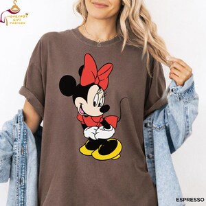 May include: Espresso-colored t-shirt featuring a cartoon Minnie Mouse design. Minnie Mouse is depicted in her classic outfit with a red bow, dress, and yellow shoes. The shirt has a relaxed fit and rolled-up sleeves.