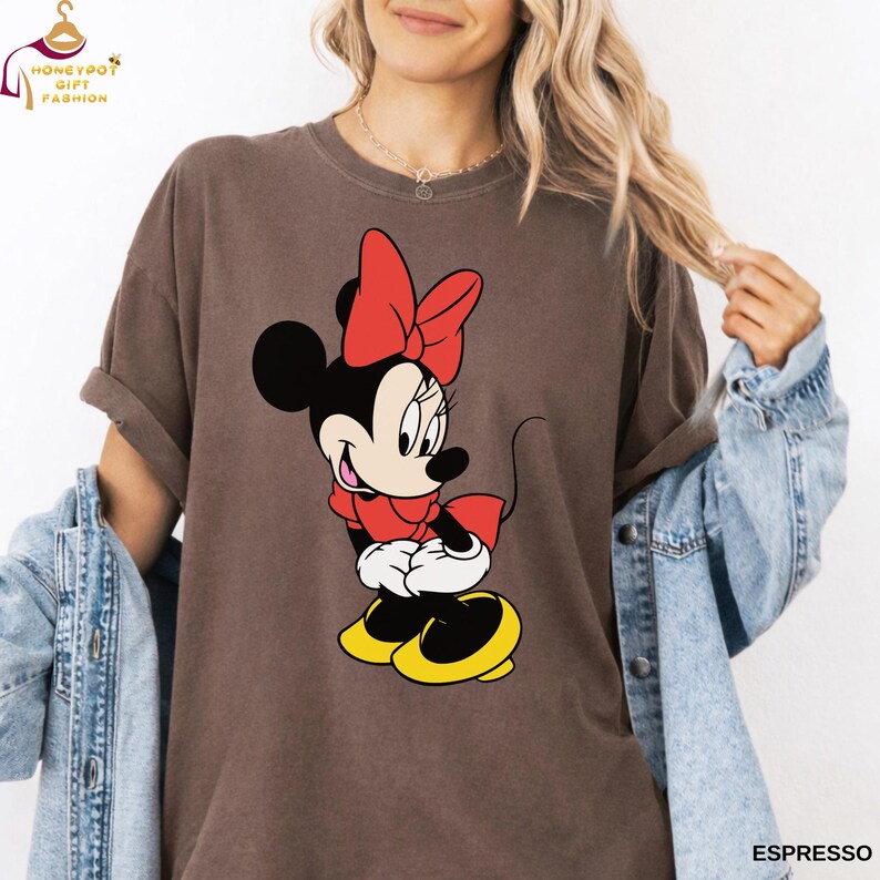 May include: Espresso-colored t-shirt featuring a cartoon Minnie Mouse design. Minnie Mouse is depicted in her classic outfit with a red bow, dress, and yellow shoes. The shirt has a relaxed fit and rolled-up sleeves.