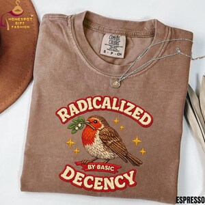 May include: Espresso-colored t-shirt with the embroidered text "RADICALIZED BY BASIC DECENCY" above and below a detailed bird illustration. The bird holds a sprig in its beak, and gold stars surround it. A silver necklace with a pendant rests on the shirt.