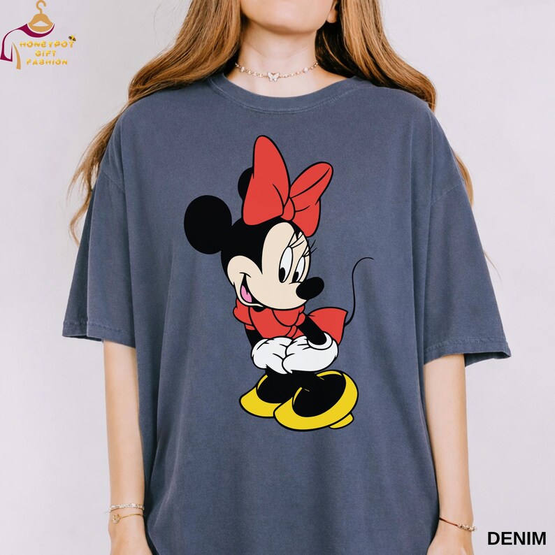 May include: Denim-colored t-shirt featuring a graphic of Minnie Mouse in a red dress and bow, white gloves, and yellow shoes. The shirt has short sleeves and a relaxed fit. The word "DENIM" is printed in white on the bottom right.