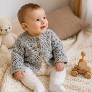 May include: A light gray long-sleeved crochet cardigan featuring a round neckline and three small brown wooden buttons down the front. The garment has a textured, open-work stitch pattern on the sleeves and body, providing a lightweight appearance.