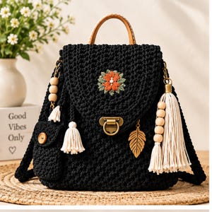 May include: A black crocheted backpack featuring a tan leather top handle, a central orange floral embroidery, and a brass-toned metal clasp. The bag is adorned with white tassels, wooden beads, and a gold-toned metal leaf charm on the side.