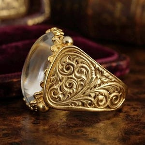 Puede incluir: Un anillo de oro con una gran piedra cabujón ovalada, lisa y translúcida, montada en una banda con intrincados patrones de volutas y filigrana en relieve que cubren los lados. El metal tiene un acabado dorado cálido y pulido.