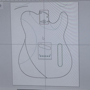 May include: A digital outline of a guitar body, including the headstock, bridge, and pickup cavities. The design is shown on a computer screen, with the file name "gravity guitar.svg" displayed.