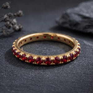 May include: A gold eternity band ring featuring a continuous row of round-cut, deep red gemstones set in a prong setting. The metal band has a polished finish, and the stones are uniform in size and color, encircling the entire circumference.