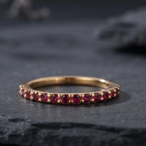 Op de afbeelding: Een dunne gouden ring met een doorlopende rij kleine, ronde, dieprode edelstenen die langs de voorste helft van de band zijn gezet. Het metaal heeft een gepolijste afwerking en de stenen zijn uniform in grootte en kleur voor een strak ontwerp.
