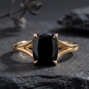 May include: A gold ring featuring a large, cushion-cut black gemstone set in a four-prong mounting. The band has a split-shank design that tapers toward the center stone, creating a sleek and polished appearance with a warm metallic finish.