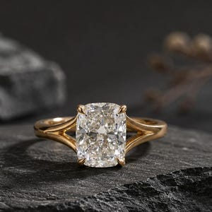 May include: A gold ring featuring a large, clear, cushion-cut center stone. The band has a split-shank design that tapers toward the stone, creating a sleek, polished look. The metal has a warm, yellow-gold tone that contrasts with the stone.