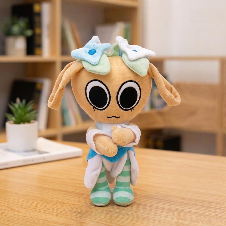 Pu&ograve; includere: Una bambola di peluche con testa beige, grandi occhi neri e lunghe orecchie flosce. La bambola indossa un completo blu e bianco con gambe a righe verdi e bianche. &Egrave; decorata con elementi a forma di stella blu e bianca sulla testa.