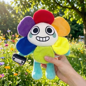 Pode incluir: Um brinquedo de pel&uacute;cia em forma de flor com um rosto sorridente e p&eacute;talas coloridas em vermelho, laranja, amarelo, verde, azul e roxo. O brinquedo est&aacute; segurando uma pequena m&aacute;scara preta. O fundo &eacute; um campo de flores.