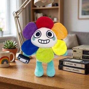Pode incluir: Um brinquedo de pel&uacute;cia em forma de flor com um rosto sorridente, p&eacute;talas de arco-&iacute;ris e pernas turquesa. O brinquedo est&aacute; segurando uma pequena fita VHS. Uma caneca com um desenho de arco-&iacute;ris e duas fitas VHS com os r&oacute;tulos "Summer '98" e "Family Movies" tamb&eacute;m s&atilde;o vis&iacute;veis.