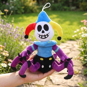May include: A plush toy in the shape of a jester with a skull face, wearing a blue, red, and yellow hat. The toy has purple arms and legs, and a black body. It has a white string for hanging.