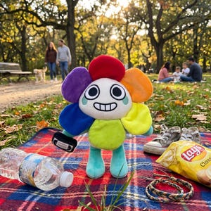 Pode incluir: Um brinquedo de pel&uacute;cia colorido em forma de flor com um rosto sorridente, segurando um pequeno objeto preto, est&aacute; sobre uma manta xadrez vermelha e azul. Uma garrafa de &aacute;gua, um saco de batatas fritas e um par de t&ecirc;nis tamb&eacute;m est&atilde;o na manta.