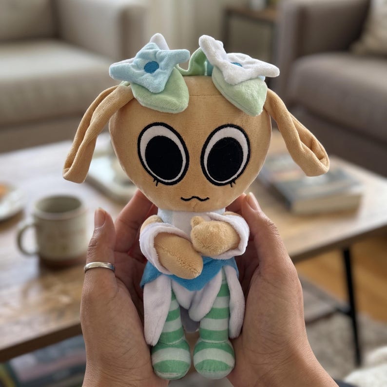 Pu&ograve; includere: Una bambola di peluche con un viso marrone chiaro, grandi occhi neri e una bocca piccola e curva. La bambola ha lunghe orecchie marroni chiare ed &egrave; adornata con elementi a forma di fiore blu e bianco. Indossa un completo blu e bianco con gambe a righe verdi e bianche.