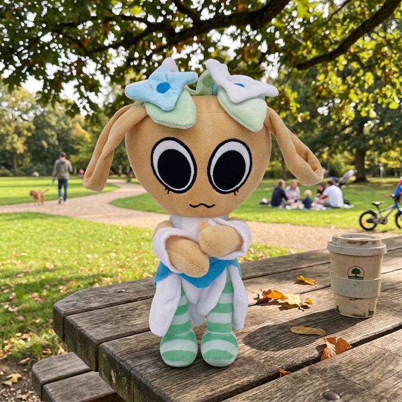 Pu&ograve; includere: Una bambola di peluche con la testa color beige, grandi occhi neri e lunghe orecchie flosce. Indossa un completo blu e bianco con gambe a righe verdi e bianche. La bambola &egrave; su un tavolo da picnic di legno in un parco.