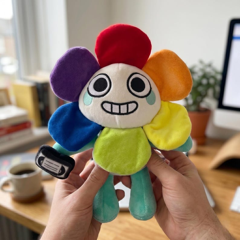 Pode incluir: Um brinquedo de pel&uacute;cia em forma de flor com um rosto sorridente e p&eacute;talas coloridas em vermelho, roxo, azul, laranja e amarelo. O brinquedo tem um caule e pernas verdes e segura uma pequena m&aacute;scara em preto e branco. O brinquedo &eacute; segurado por duas m&atilde;os.