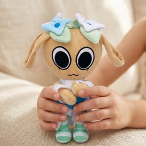 Pu&ograve; includere: Una bambola di peluche con una testa color beige, grandi occhi neri e una bocca piccola e curva. La bambola ha lunghe orecchie flosce ed &egrave; adornata da elementi a forma di fiore blu e bianco. Indossa un completo blu e bianco con piedi a righe verdi e bianche.