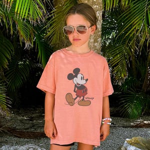 May include: A coral-colored t-shirt featuring a graphic of Mickey Mouse. The cartoon character is depicted in his classic pose, with a red shorts, brown shoes, and white gloves. The word "Disney" is printed below the image. The shirt is worn by a person wearing sunglasses.