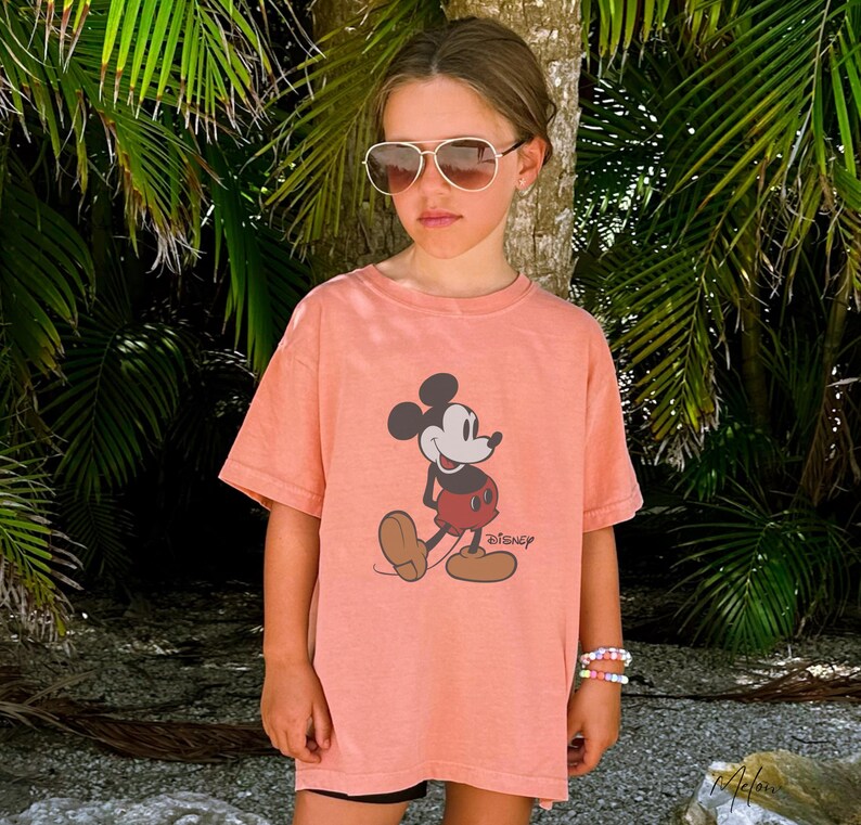 May include: A coral-colored t-shirt featuring a graphic of Mickey Mouse. The cartoon character is depicted in his classic pose, with a red shorts, brown shoes, and white gloves. The word "Disney" is printed below the image. The shirt is worn by a person wearing sunglasses.
