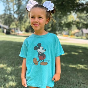May include: A turquoise t-shirt featuring a classic Mickey Mouse graphic. The cartoon character is depicted in black, red, and brown, with the Disney logo below. The shirt is worn by a person with white bows in their hair.