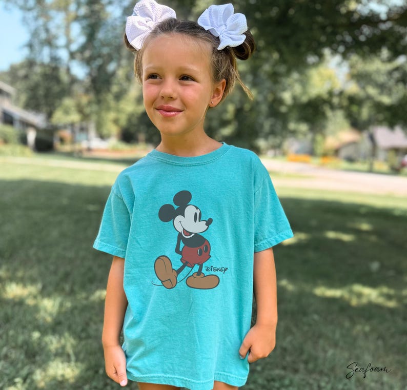 May include: A turquoise t-shirt featuring a classic Mickey Mouse graphic. The cartoon character is depicted in black, red, and brown, with the Disney logo below. The shirt is worn by a person with white bows in their hair.