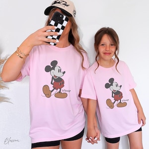 May include: Two light pink t-shirts featuring a Mickey Mouse graphic with the Disney logo. The cartoon character is in black, red, and brown. The shirts are worn by two people, one wearing a baseball cap and holding a phone.