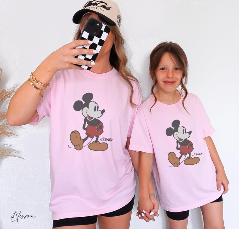 May include: Two light pink t-shirts featuring a Mickey Mouse graphic with the Disney logo. The cartoon character is in black, red, and brown. The shirts are worn by two people, one wearing a baseball cap and holding a phone.