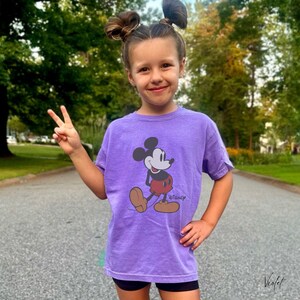 May include: A purple t-shirt featuring a graphic of Mickey Mouse in a walking pose. The Disney logo is printed below the character. The shirt is worn by a person who is giving a peace sign.