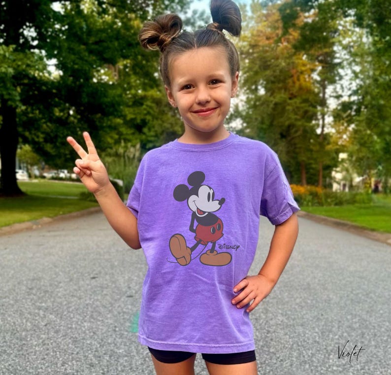 May include: A purple t-shirt featuring a graphic of Mickey Mouse in a walking pose. The Disney logo is printed below the character. The shirt is worn by a person who is giving a peace sign.