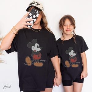 May include: Black t-shirts featuring a classic Mickey Mouse design. The cartoon character is depicted in a walking pose with red shorts, brown shoes, and a gray outline. The word "Disney" is printed below the character. The word "Black" is printed at the bottom left.