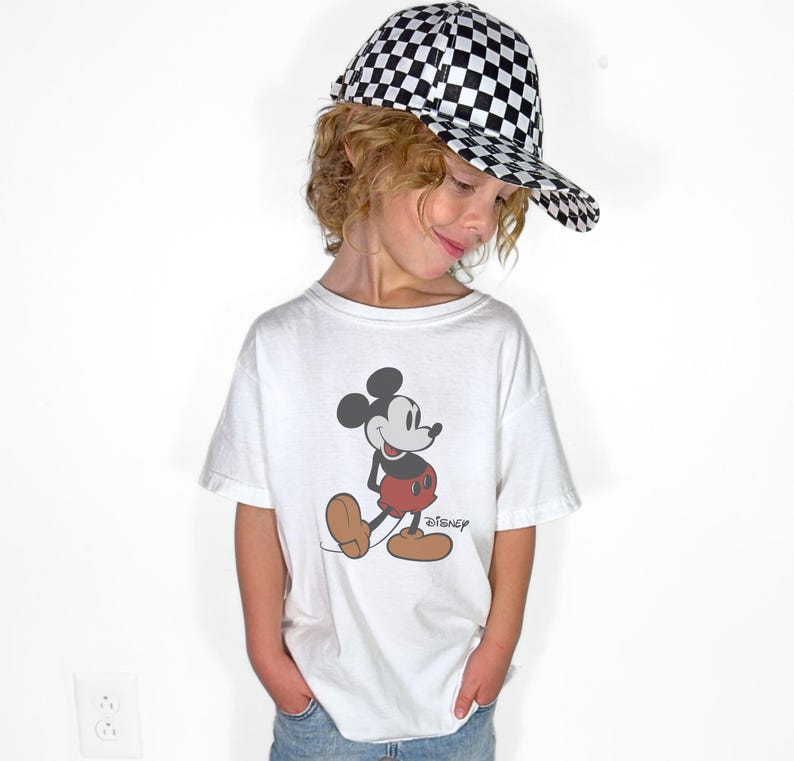 May include: A white t-shirt featuring a Mickey Mouse graphic, paired with a black and white checkered baseball cap. The Disney logo is visible on the shirt. The outfit is worn by a person.