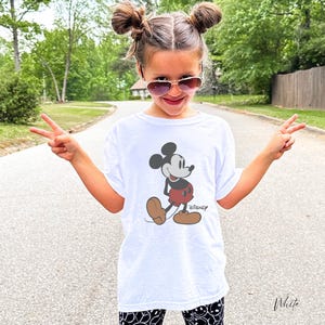 May include: White t-shirt featuring a classic Mickey Mouse graphic. The cartoon character is depicted in black, red, and brown. The word "Disney" is printed below the character. The shirt is worn by a person.