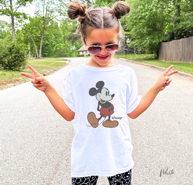 May include: White t-shirt featuring a classic Mickey Mouse graphic. The cartoon character is depicted in black, red, and brown. The word "Disney" is printed below the character. The shirt is worn by a person.