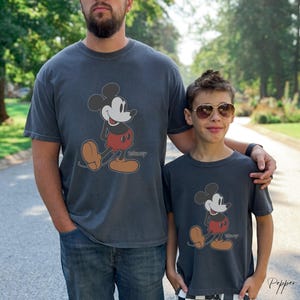 May include: Two charcoal gray t-shirts featuring a classic Mickey Mouse graphic. The cartoon character is depicted in black, red, and tan colors. The word "Disney" is printed below the character. The shirts are worn by two people.