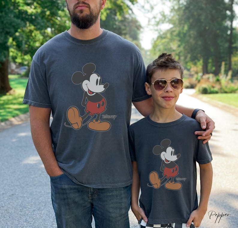 May include: Two charcoal gray t-shirts featuring a classic Mickey Mouse graphic. The cartoon character is depicted in black, red, and tan colors. The word "Disney" is printed below the character. The shirts are worn by two people.