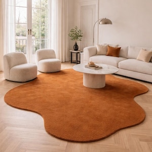 Op de afbeelding: Een oranje, organisch gevormd vloerkleed in een woonkamer. Het kleed ligt op een lichte houten vloer en wordt omringd door crèmekleurige meubels, waaronder een bank en twee fauteuils. Een witte salontafel staat op het kleed.