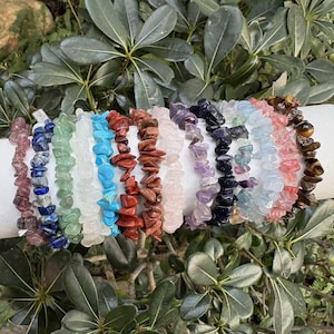 May include: A collection of gemstone chip bracelets in various colors, including pink, blue, green, and brown. The bracelets are displayed on a white cylinder with a gold band, surrounded by green foliage.