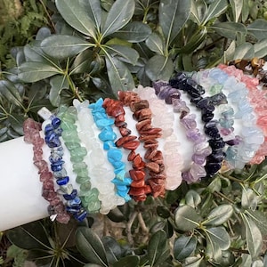Può includere: Una collezione di braccialetti con chip di pietre preziose in vari colori, tra cui blu, verde, rosso e viola. I braccialetti sono esposti su un oggetto cilindrico bianco, con fogliame verde sullo sfondo. Ogni braccialetto è fatto di piccole pietre di forma irregolare.