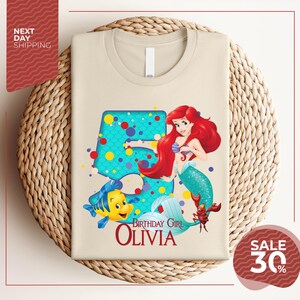A t-shirt featuring a design of the Disney character Ariel from The Little Mermaid, with the text "Birthday Girl Olivia" and a price of $30.