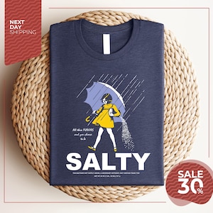 a blue t-shirt with a graphic design depicting a person holding an umbrella and sprinkling salt on the ground. The text on the t-shirt reads "All these flavors and to choose one, you must be salty".