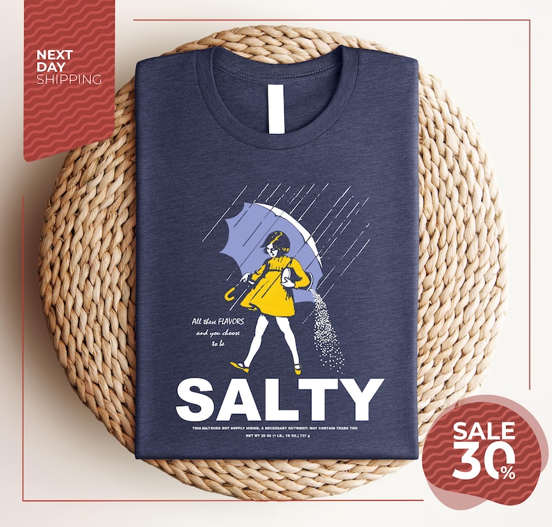 a blue t-shirt with a graphic design depicting a person holding an umbrella and sprinkling salt on the ground. The text on the t-shirt reads "All these flavors and to choose one, you must be salty".