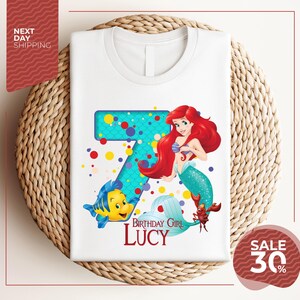 A white t-shirt with a design featuring the character Ariel from the Disney movie "The Little Mermaid". The design includes a large number 7 and a mermaid character, along with the text "Birthday Girl Lucy".