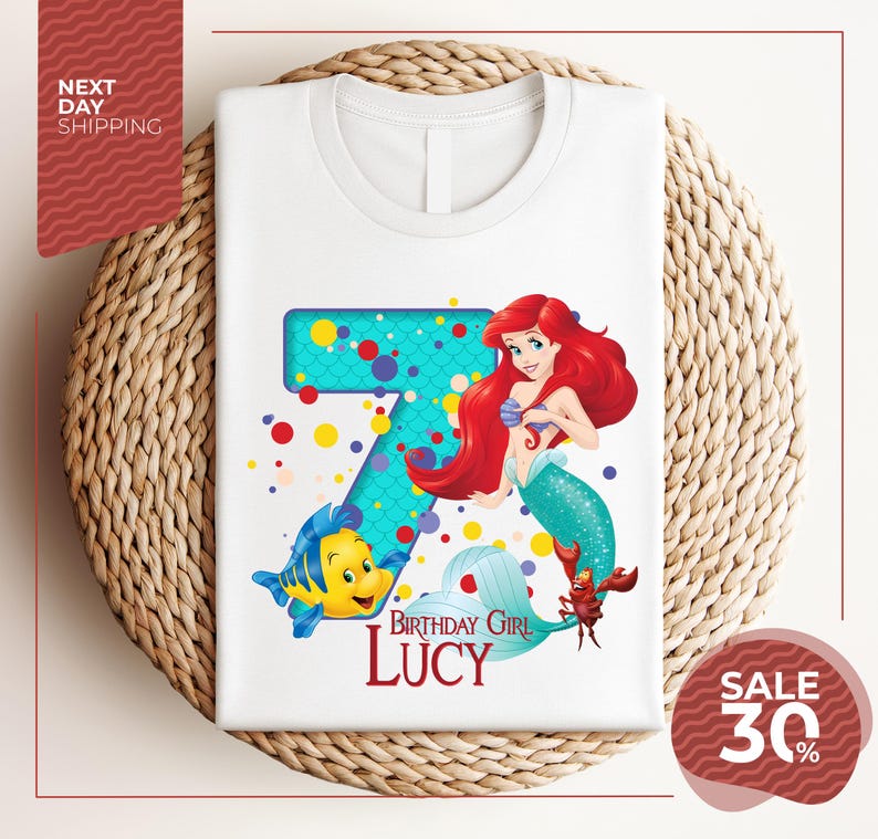 A white t-shirt with a design featuring the character Ariel from the Disney movie "The Little Mermaid". The design includes a large number 7 and a mermaid character, along with the text "Birthday Girl Lucy".