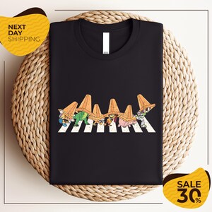 a black t-shirt with a graphic design of cartoon characters wearing sombreros, arranged in a crosswalk pattern. The t-shirt is displayed on a woven placemat, and there are two yellow sale signs in the image.